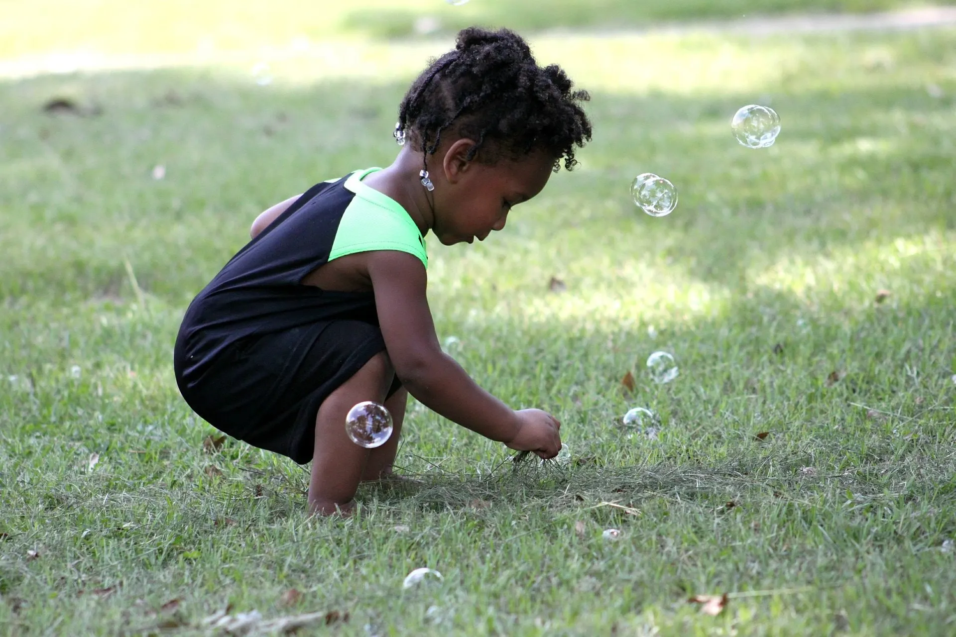 A young child crouching in the grass reaching for soap bubbles